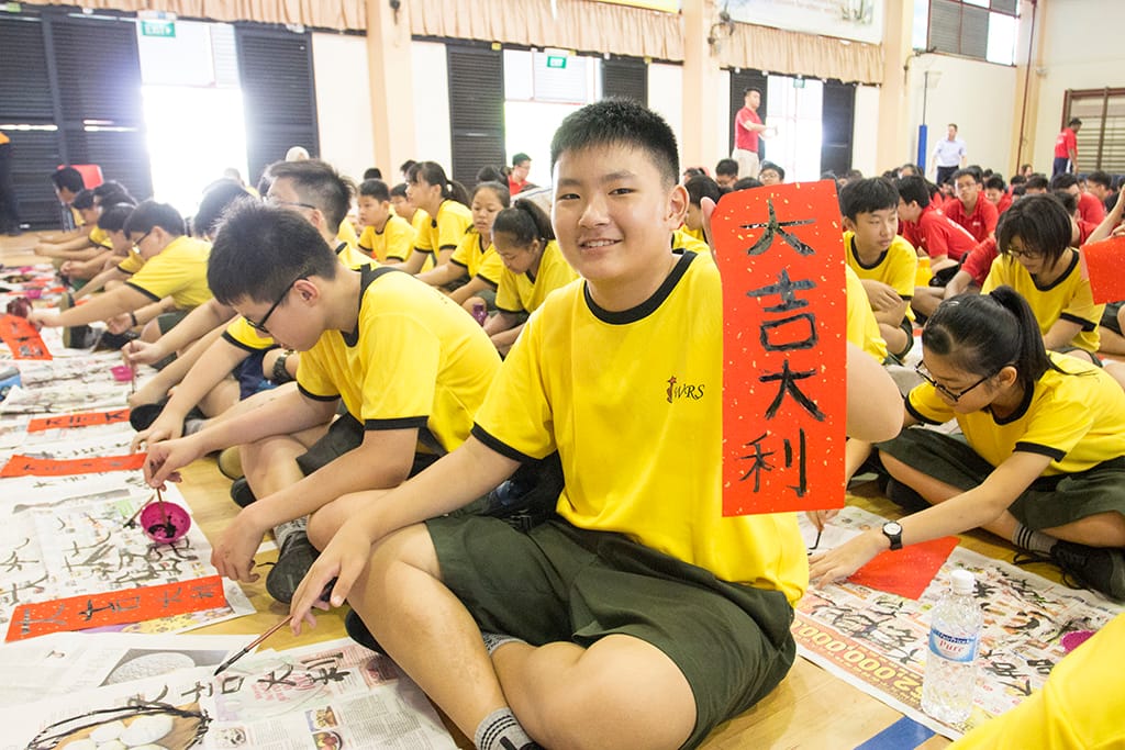 A smiling boy in a yellow shirt holds a red card with Chinese calligraphy while seated on a gym floor with other students.